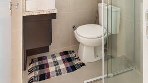 A modern bathroom with a glass shower enclosure, a toilet, a sink with a towel rack above, and a small shelf with toiletries. - Studio c/ churrasq 650m da Torre Panorâmica RME047 (Curitiba)