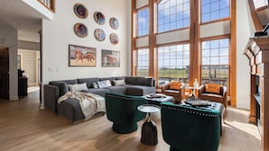 Ein großzügiges Wohnzimmer mit einer großen Eckcouch, einem Couchtisch und einem Kamin. Große Fenster bieten einen Blick ins Freie.