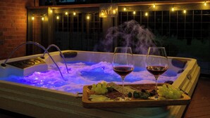 A hot tub with a wooden tray of grapes and two glasses of wine, surrounded by string lights and a 'BATHROOM' sign.