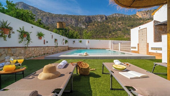 A poolside area with lounge chairs, a table with drinks, and a mountain backdrop.