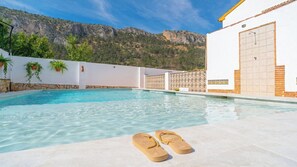 A swimming pool with a pair of sandals, a white building with a tiled wall, and a mountainous backdrop.