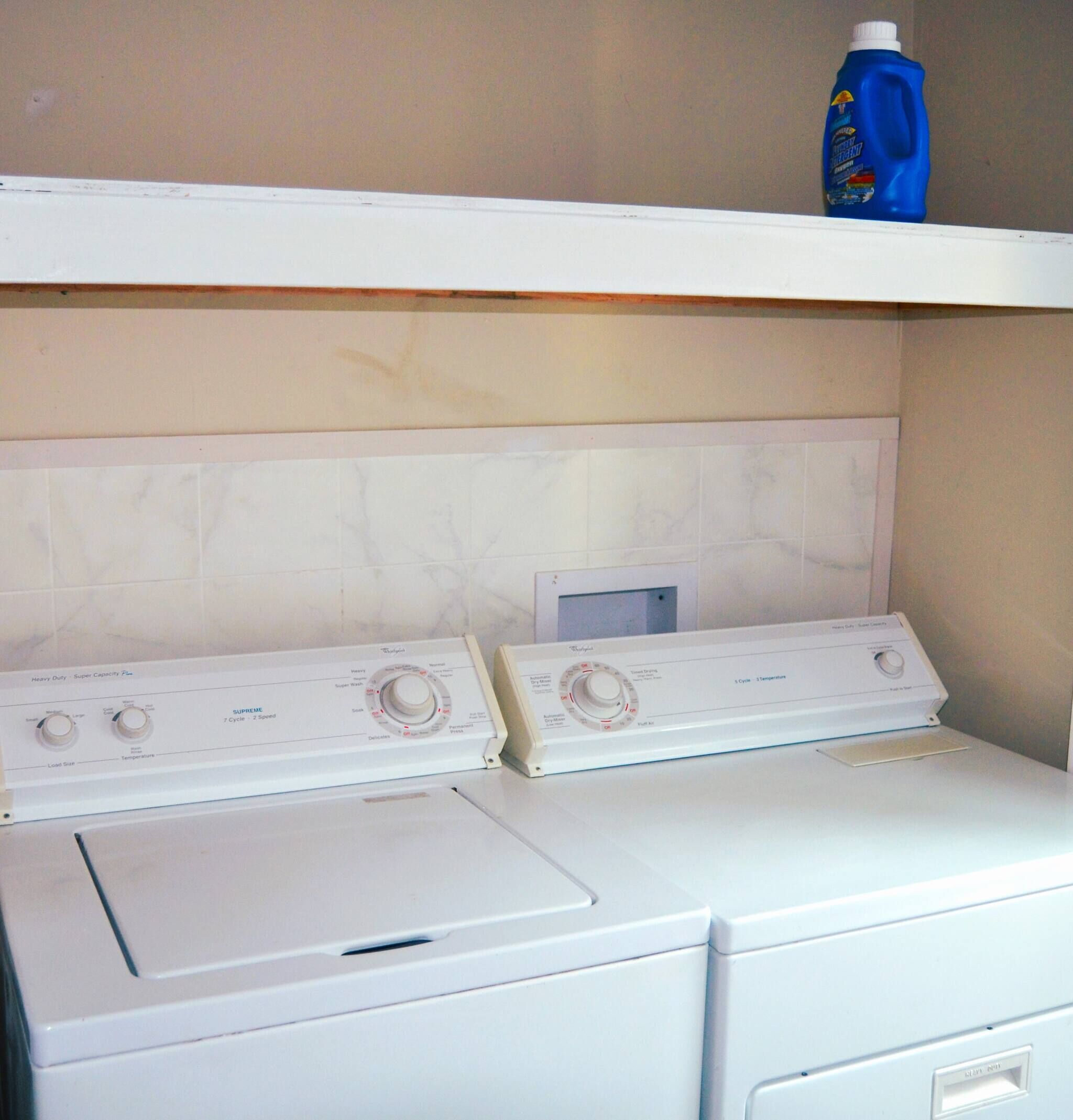 A front-loading washer and dryer unit with control knobs and a detergent bottle on a shelf above.