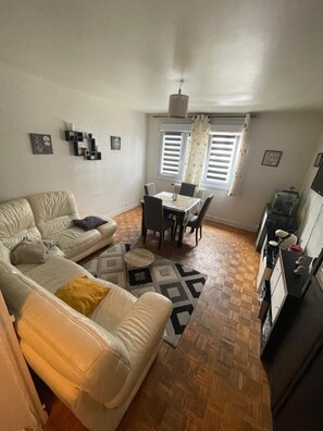 A living room with a sofa, a dining table with chairs, a kitchen area, and a window with curtains. - La Parenthèse Normande (Caen)