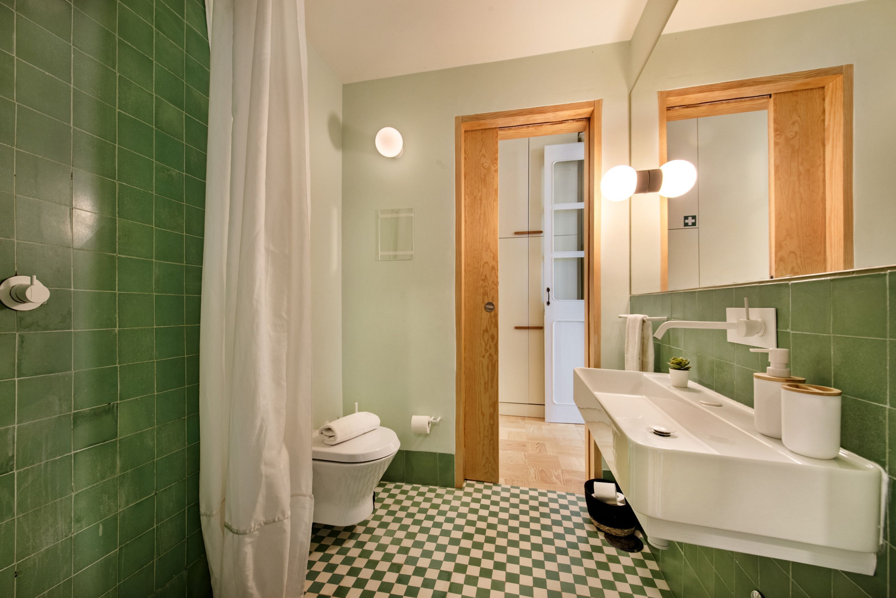 A bathroom with green tiles, a white toilet, a sink with a mirror, and a checkered floor.