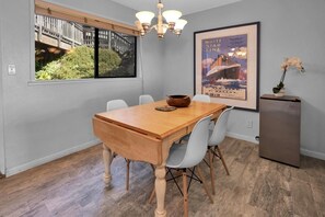 Un coin repas avec une table en bois, des chaises blanches, un lustre, une affiche encadrée et un réfrigérateur.