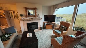Ein Wohnzimmer mit Kamin, Fernseher, Korbmöbeln und Blick ins Freie.