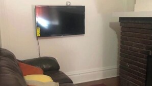 Ein Wohnzimmer mit braunem Ledersofa, einem am Wand montierten Flachbildfernseher und einem Kamin aus Backsteinen.