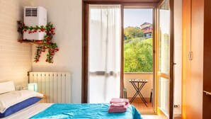 Ein Schlafzimmer mit einem Bett, einem Heizkörper, einer Klimaanlage, einem Balkon mit Tisch und Stühlen sowie Blick auf Grünflächen und ein Haus.