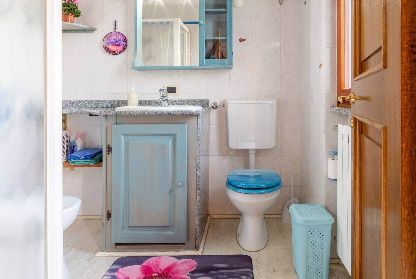 A bathroom with a blue cabinet, a white toilet with a blue seat, a small trash bin, and a mirror above the sink.