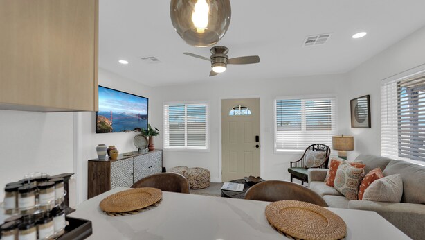 A modern living room with a white dining table, brown chairs, a sofa, and a ceiling fan.