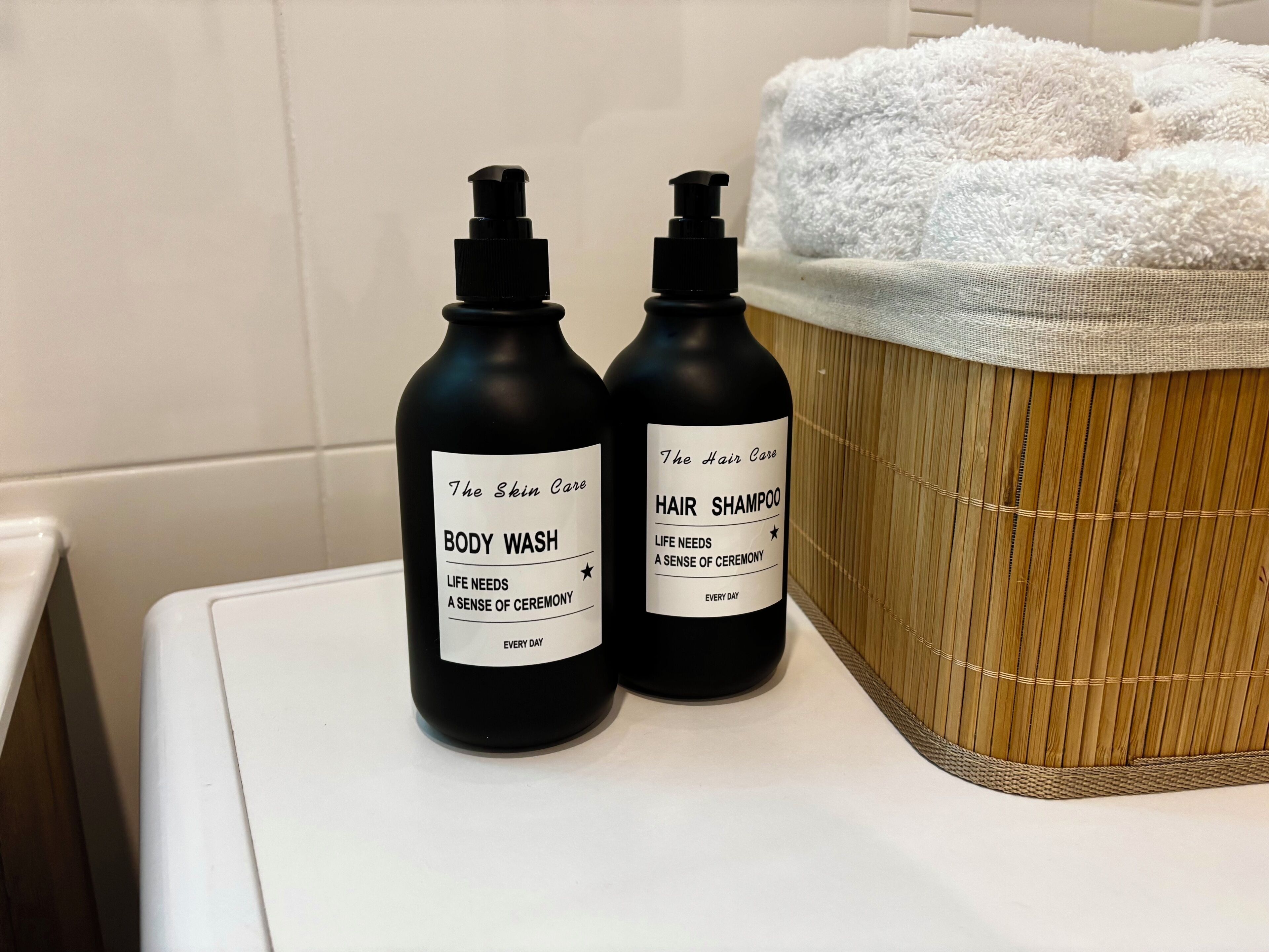 Kaksi pulloa vartalopesua ja hiusten shampoota valkoisella työtasolla, taustalla bambikori ja valkoiset pyyhkeet.