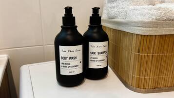Kaksi pulloa vartalopesua ja hiusten shampoota valkoisella työtasolla, taustalla bambikori ja valkoiset pyyhkeet.
