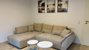 Un canapé d’angle beige, accompagné de deux petites tables rondes blanches à structure métallique, une porte blanche et trois tableaux encadrés accrochés au mur.