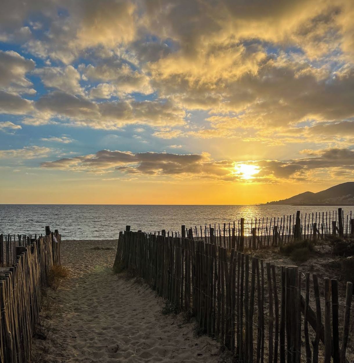 Un sendero de playa con valla de madera que lleva al mar, bajo un cielo al atardecer.