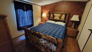 Una habitación con estructura de cama de madera, colcha a cuadros, dos lámparas de noche, una mesita de noche, una silla de madera y una ventana con cortinas.