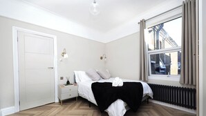 A bedroom with a bed, bedside table, lamp, window, radiator, and a view of the outside. - Newly refurbished 2 bedroom apartment in trendy Leyton (London)