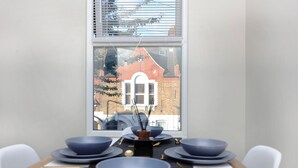 A dining area with a table set for four, a window with blinds, and a view of a brick building outside. - Newly refurbished 2 bedroom apartment in trendy Leyton (London)