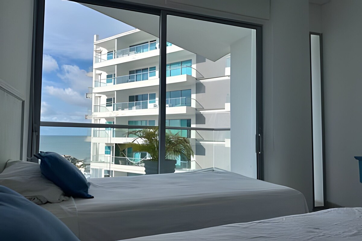 A hotel room with a large window offering a view of a modern building and the sea.