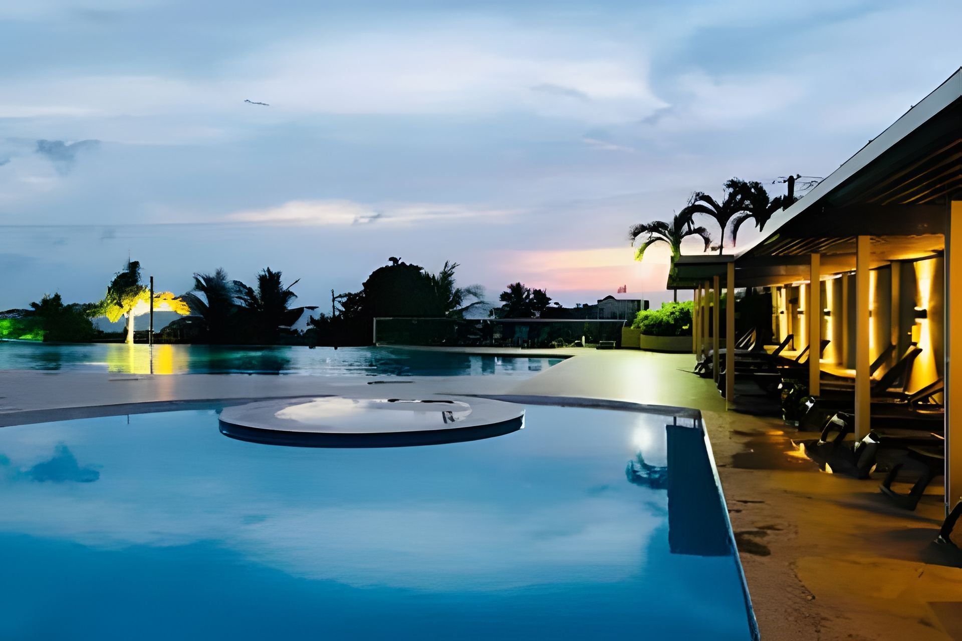 A swimming pool with a circular feature, surrounded by lounge chairs and a building with outdoor seating.