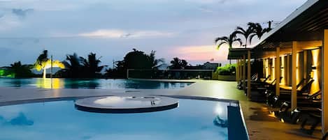 A swimming pool with a circular feature, surrounded by lounge chairs and a building with outdoor seating.