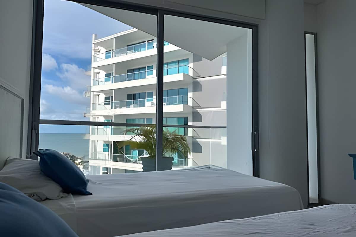 A hotel room with a large window offering a view of a modern building and the sea.