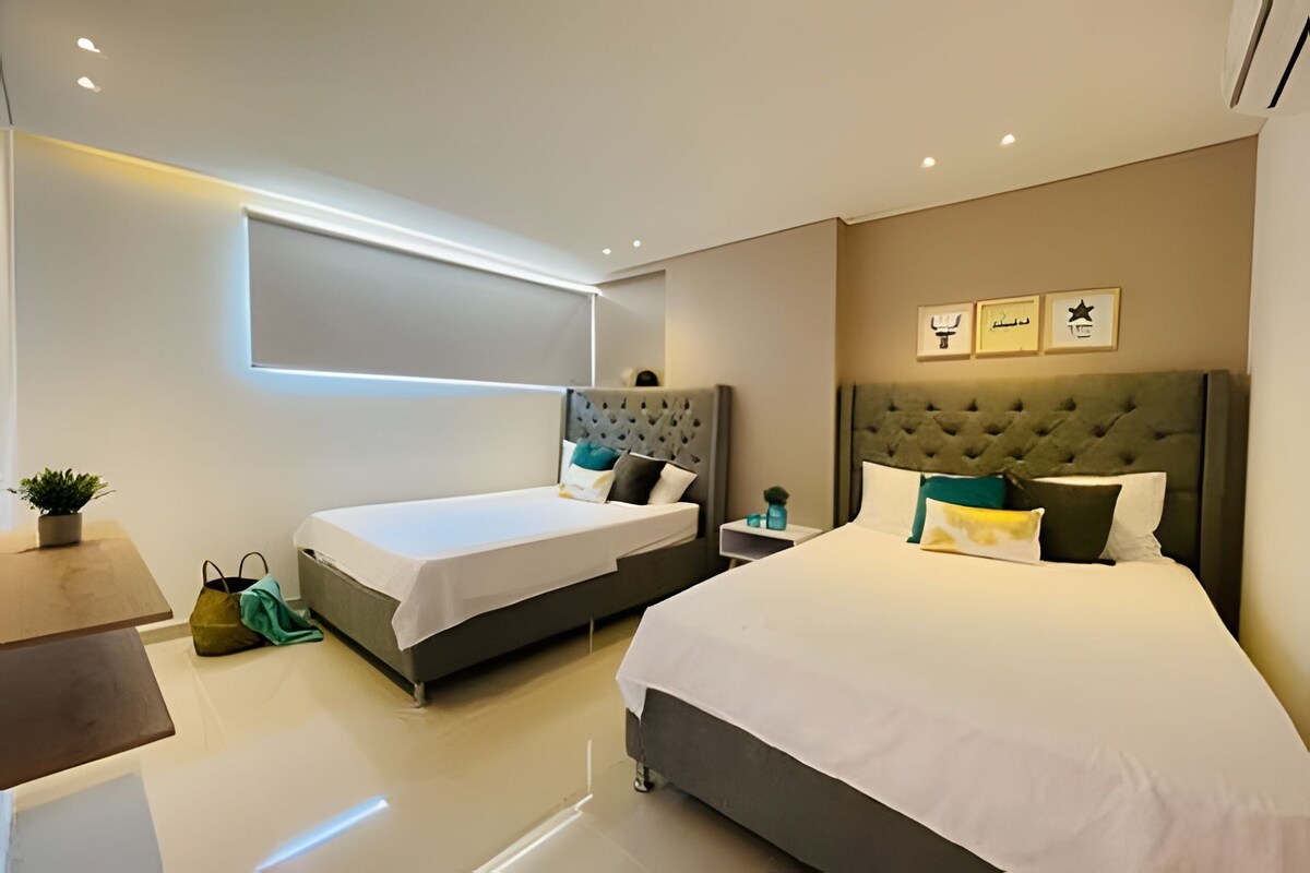 A modern hotel room with two beds, a small table, and a potted plant.