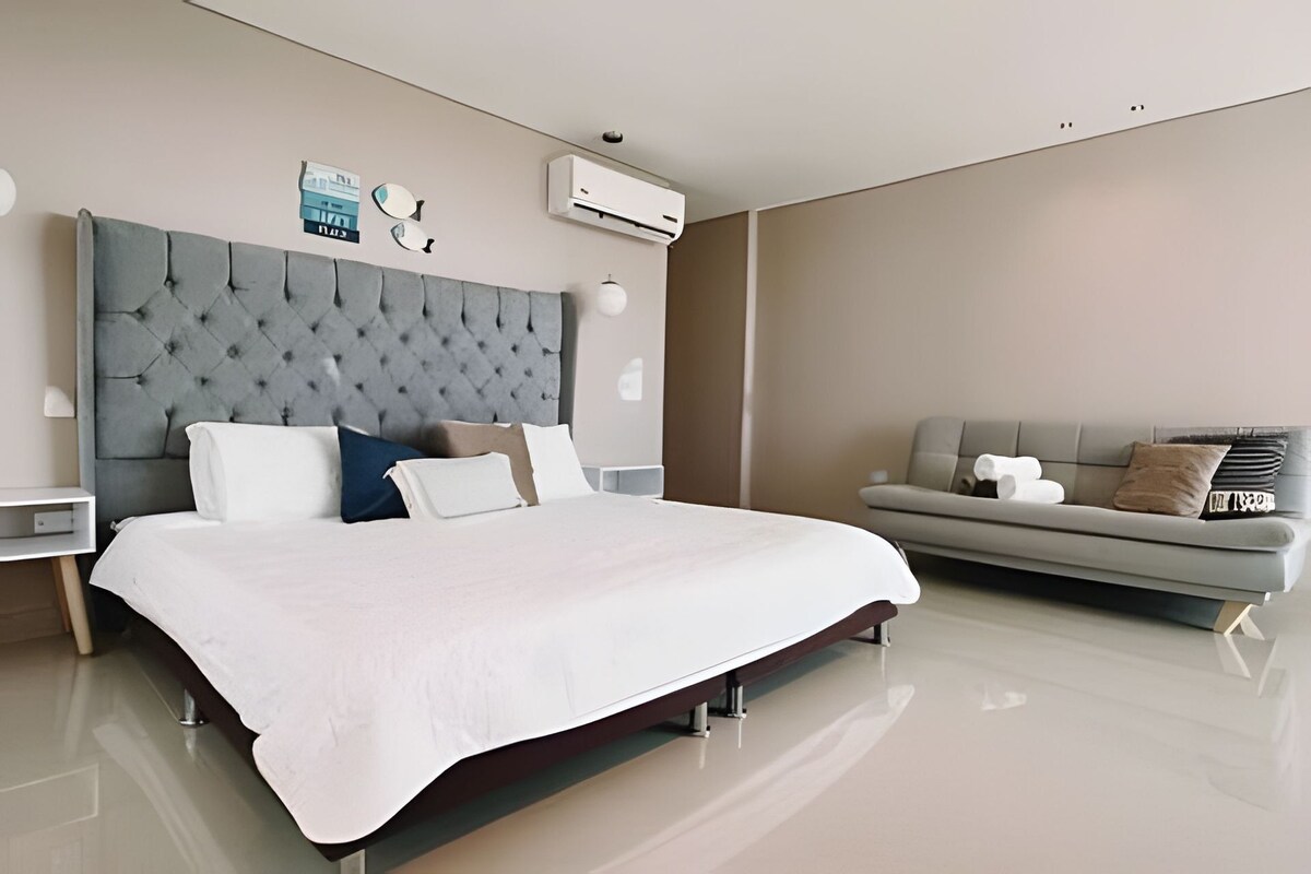 A modern bedroom with a large bed, a grey upholstered headboard, a grey sofa, and an air conditioning unit on the wall.