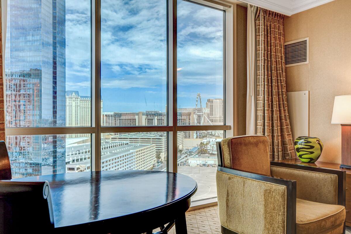 A hotel room with a large window offering a city view, a round table, two chairs, a vase, and a lamp.