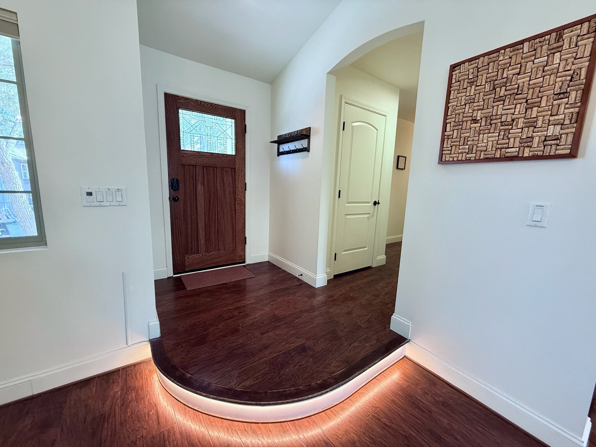 Un couloir doté d’un plancher en bois, d’une porte marron foncé et d’une porte blanche. Un luminaire est fixé au mur, et un tableau d’affichage en liège y est également accroché.