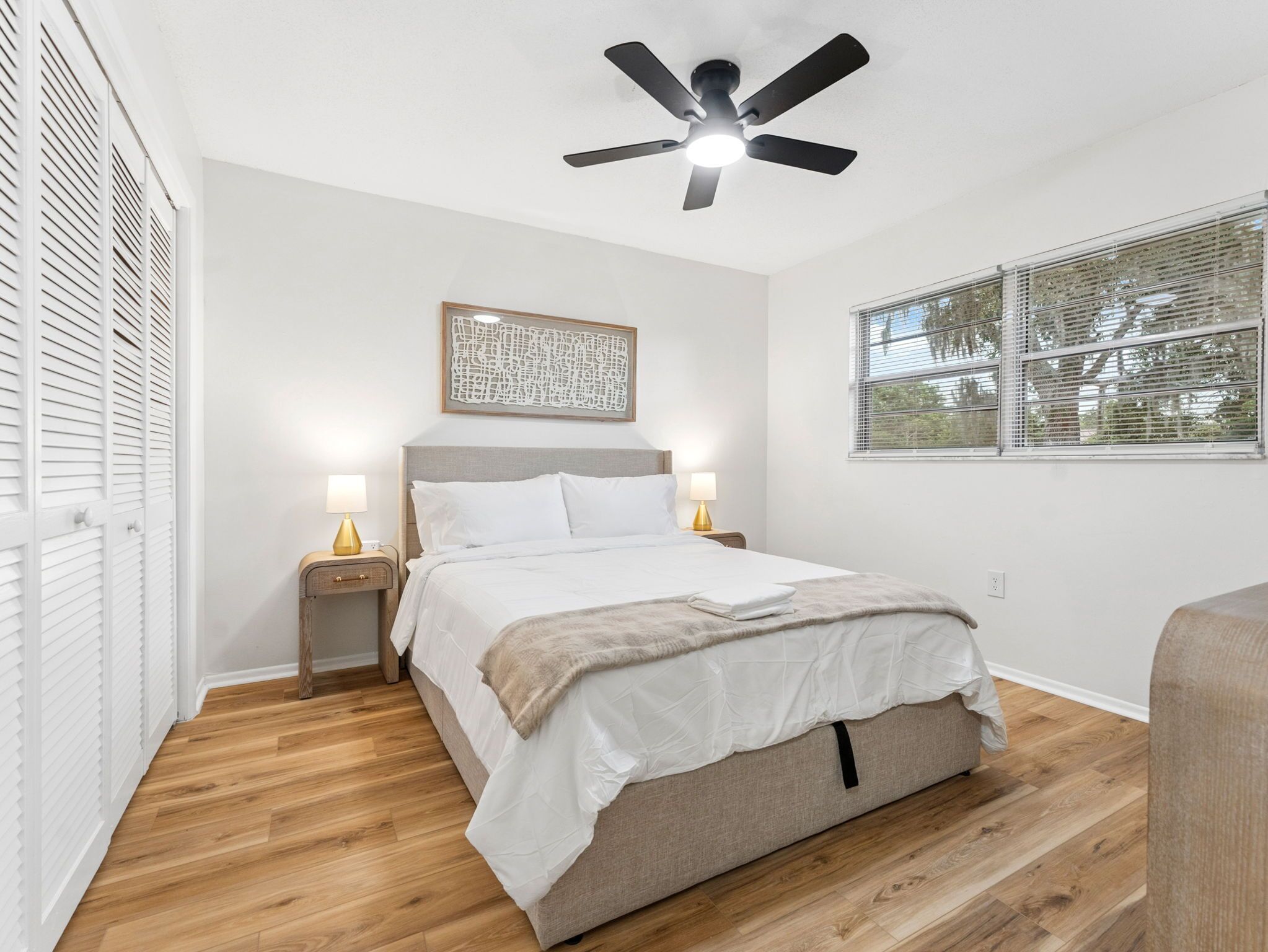 Une chambre à coucher avec un lit, des lampes de chevet, un ventilateur de plafond, une fenêtre avec des stores et un plancher en bois.
