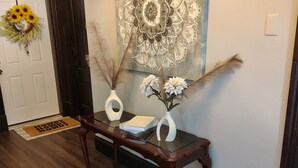 Un pasillo con piso de madera, una puerta oscura, una obra de arte colgada en la pared, un jarrón blanco con flores y un felpudo.