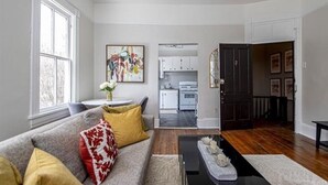Un salón con un sofá gris, una mesa de centro y un cuadro en la pared. Se ve una cocina a través de un vano abierto.