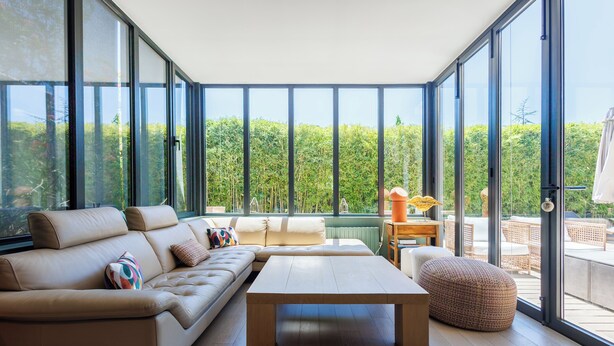 A modern living room with a sofa, coffee table, and ottoman. Large windows offer a view of greenery outside. - Villa du Val - Welkeys (Aix-en-Provence)
