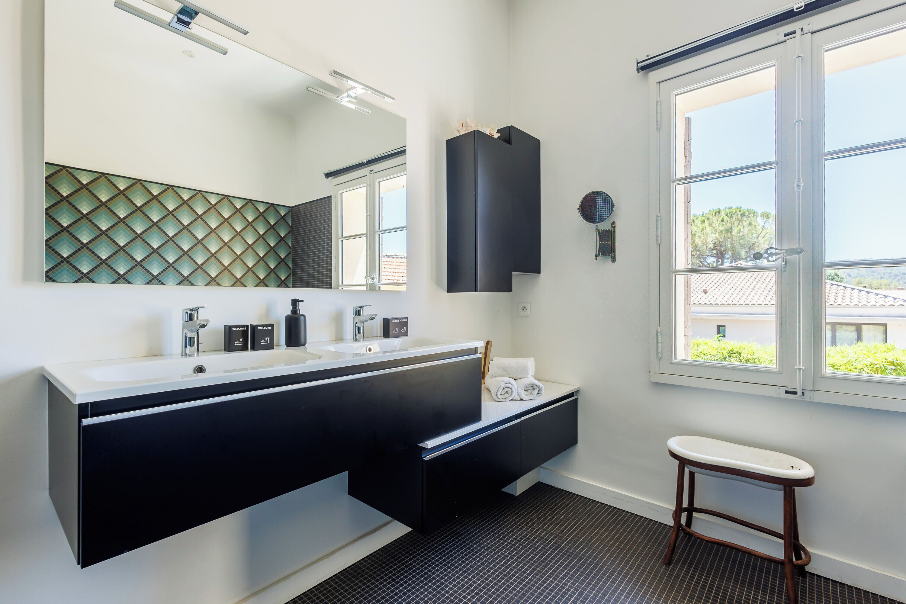 A modern bathroom with a large mirror, two sinks, a black vanity, and a window with a view of greenery.