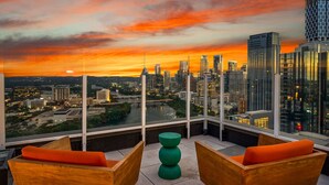 Ein Hotelzimmer in einem Hochhaus mit Blick auf die Stadt bei Sonnenuntergang, mit großem Fenster, grünem Tisch und Holzsesseln.