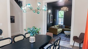 A dining area with a dark wooden table, woven chairs, and a decorative vase with green foliage. A modern chandelier with spherical glass elements hangs above.