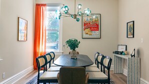 A dining room with a round table, six chairs, a vase with green plants, a vintage poster, and a window with orange curtains.