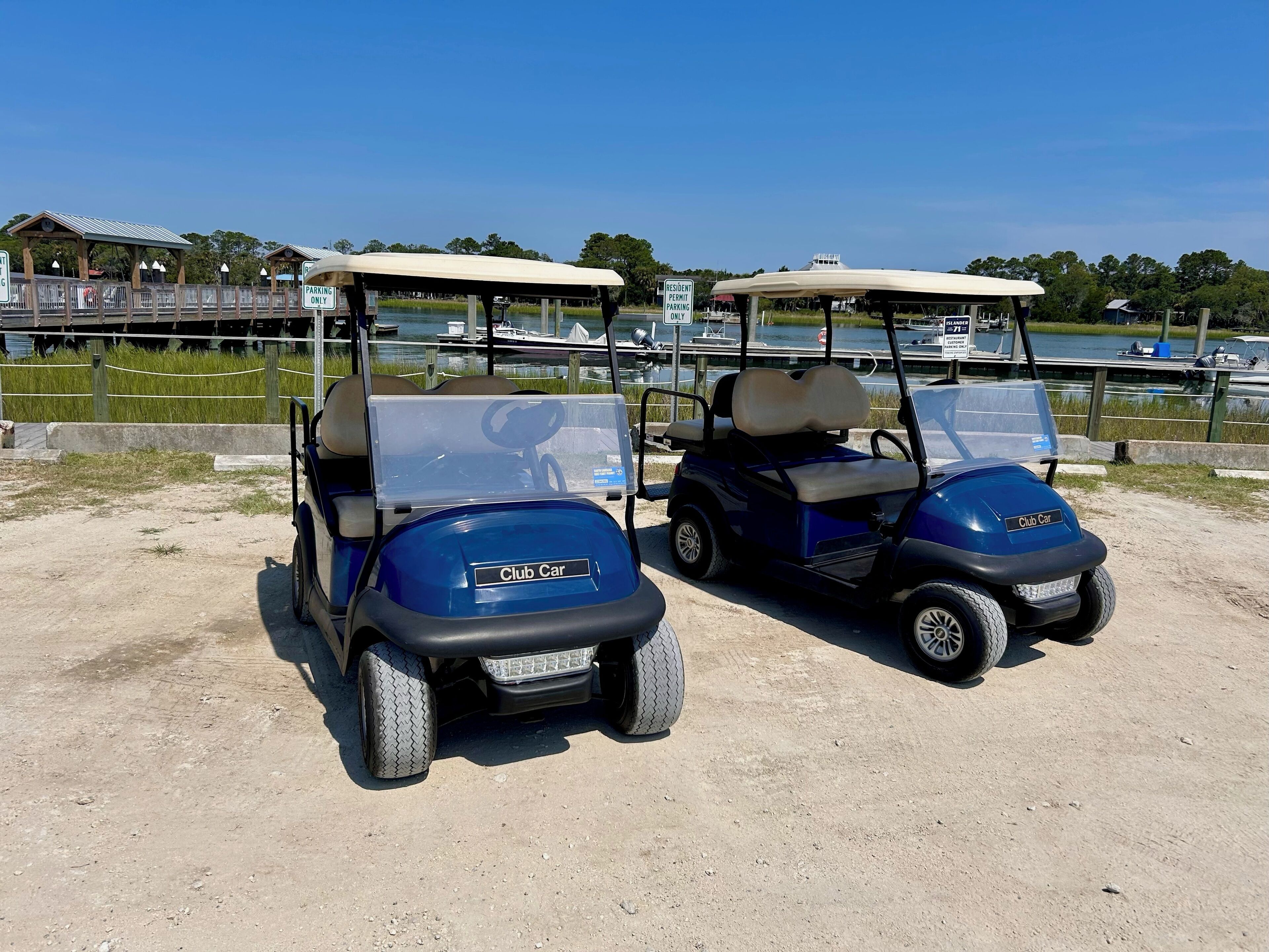 Zwei Golfcarts auf einer befestigten Fläche, im Hintergrund ein Gewässer und Boote.