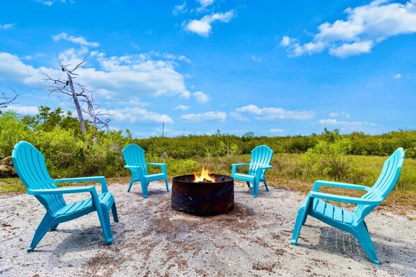 A fire pit with four turquoise Adirondack chairs arranged around it on a sandy beach with a clear sky and greenery in the background. - Ozello Waterfront Paradise (Crystal River)