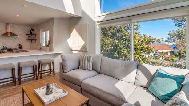 A modern living space with a kitchen area, a sofa, and a large window offering a view of greenery and buildings. - Steps to Bay & Beach | Rooftop Deck, Fire Pit, AC, Garage, Views (San Diego)