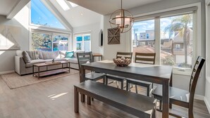 A spacious living room with a high ceiling, large windows, a dining area with a wooden table and chairs, and a cozy seating area with a sofa and cushions. - Steps to Bay & Beach | Rooftop Deck, Fire Pit, AC, Garage, Views (San Diego)
