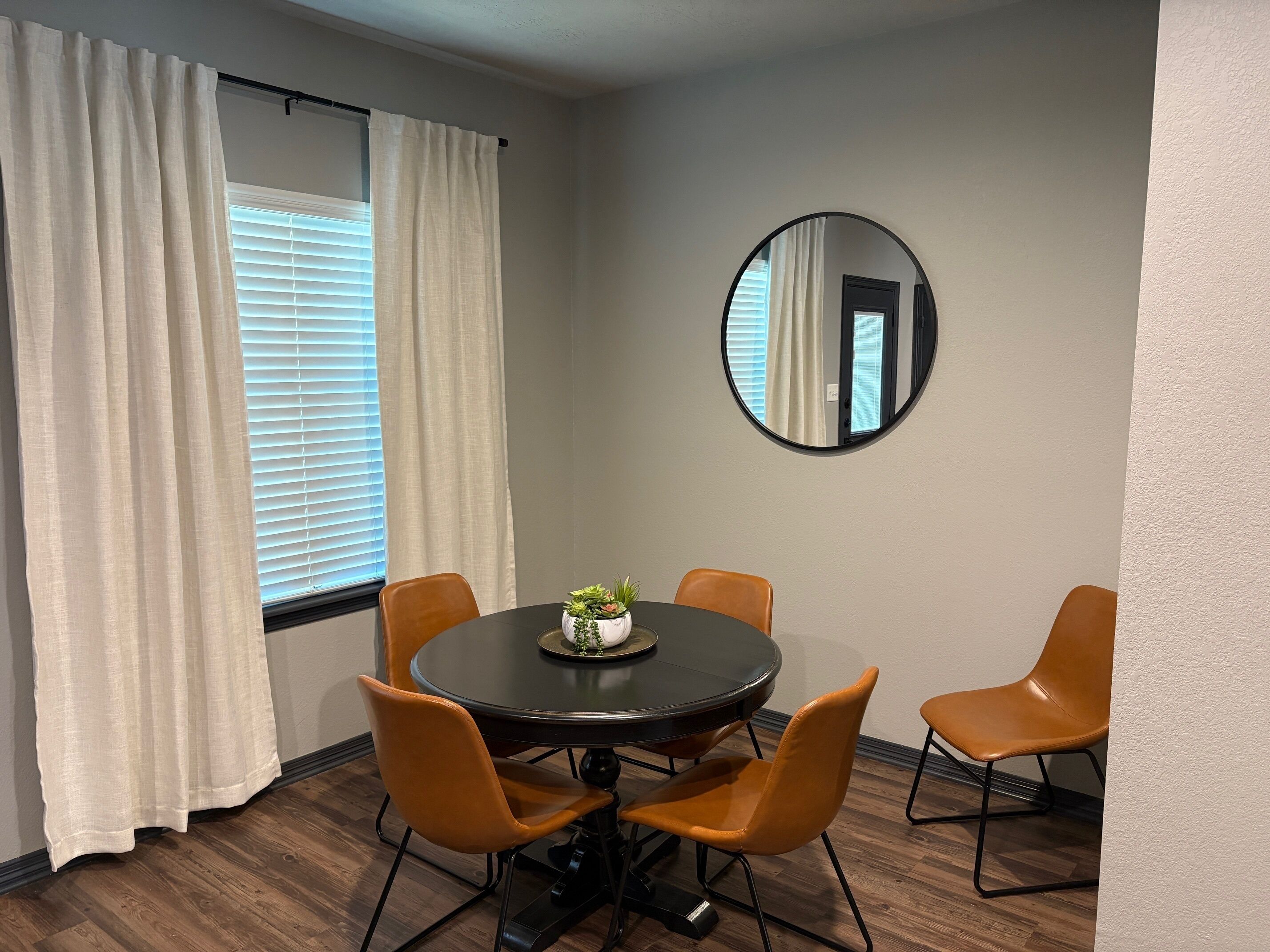 A room with a round table, four orange chairs, a potted plant, and a round mirror on the wall.