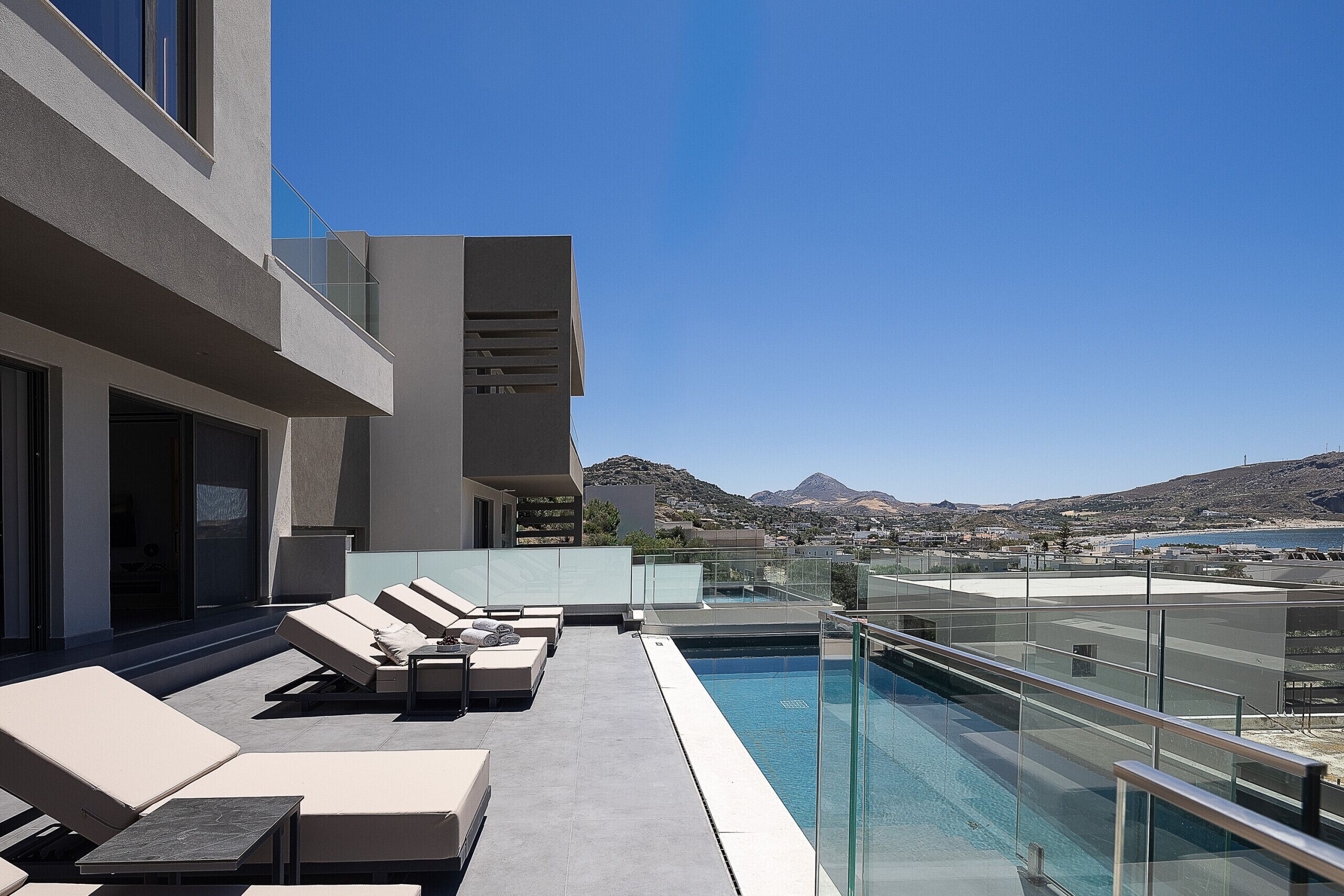 Un área de piscina en la azotea con sillones, barandilla de vidrio y vistas al mar y a las montañas.