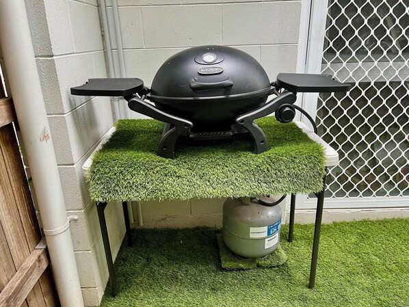 Un barbecue au gaz portatif, avec une bouteille de gaz supplémentaire, posé sur une table recouverte de gazon synthétique.