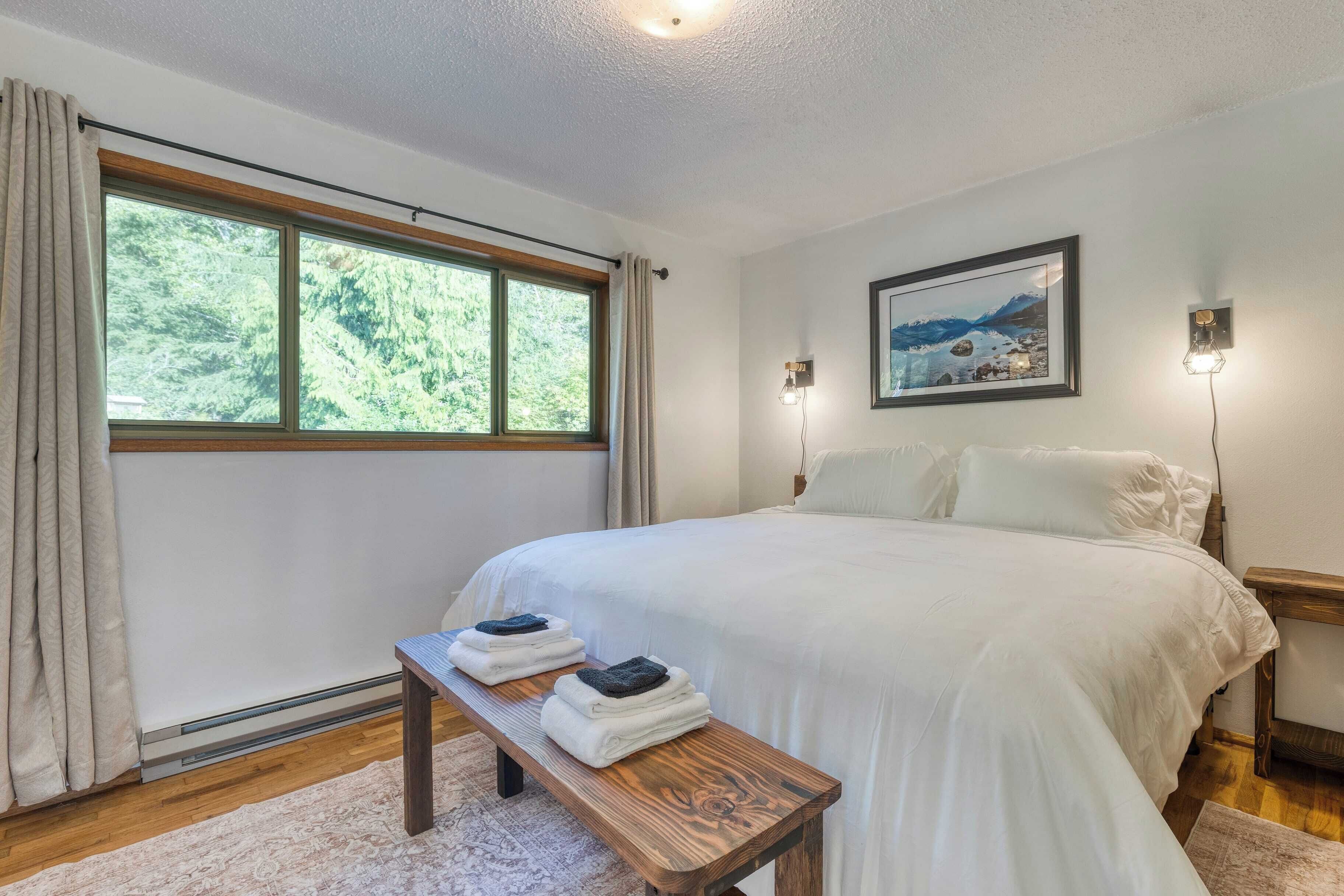 Une chambre avec un grand lit, un banc en bois avec des serviettes, une fenêtre avec des rideaux, un tableau encadré au mur et des lampes de chevet.