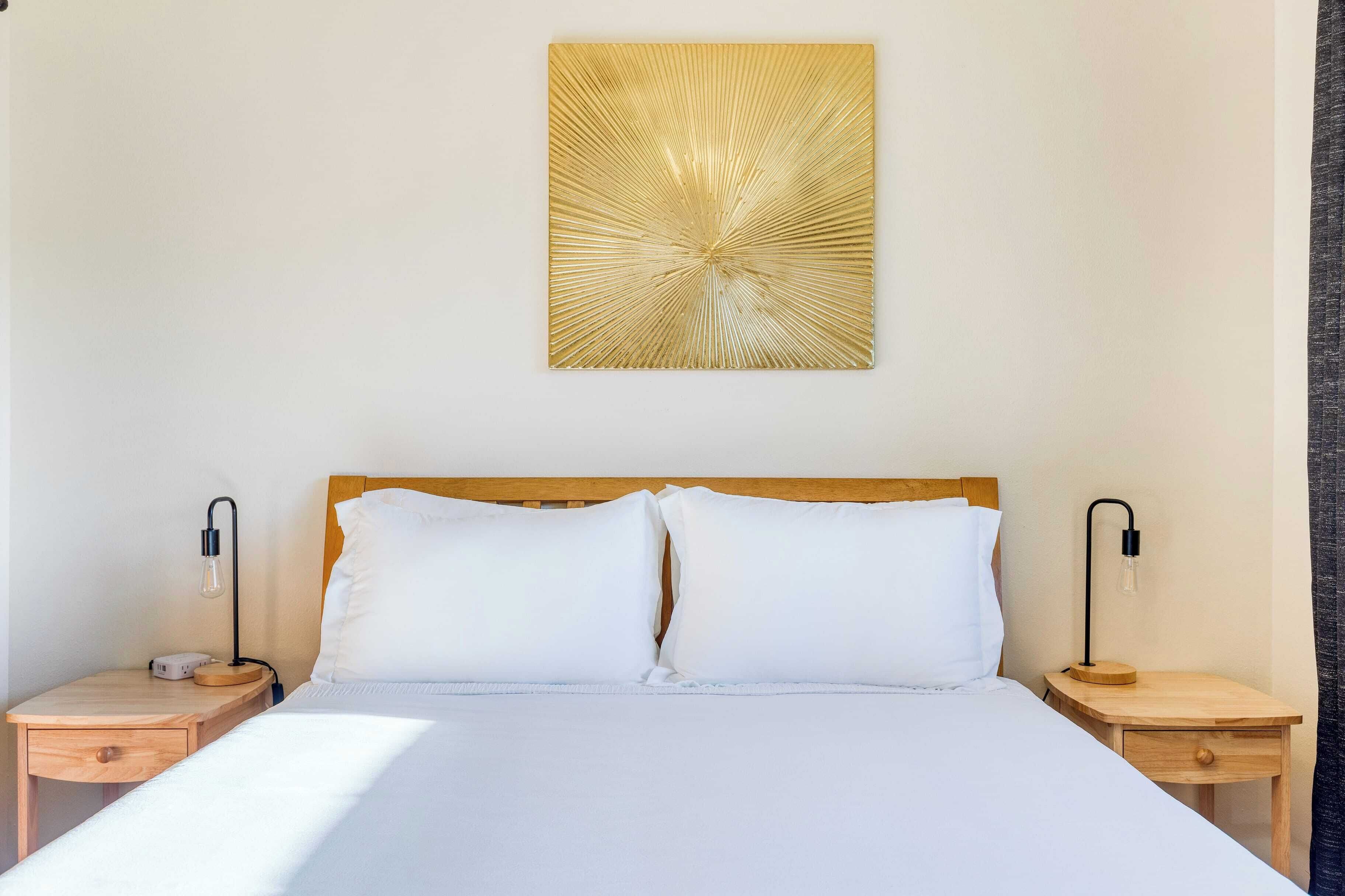 Ein ordentlich bezogenes Bett mit weißen Laken, zwei Nachttische mit Lampen und ein an der Wand hängendes Kunstwerk.
