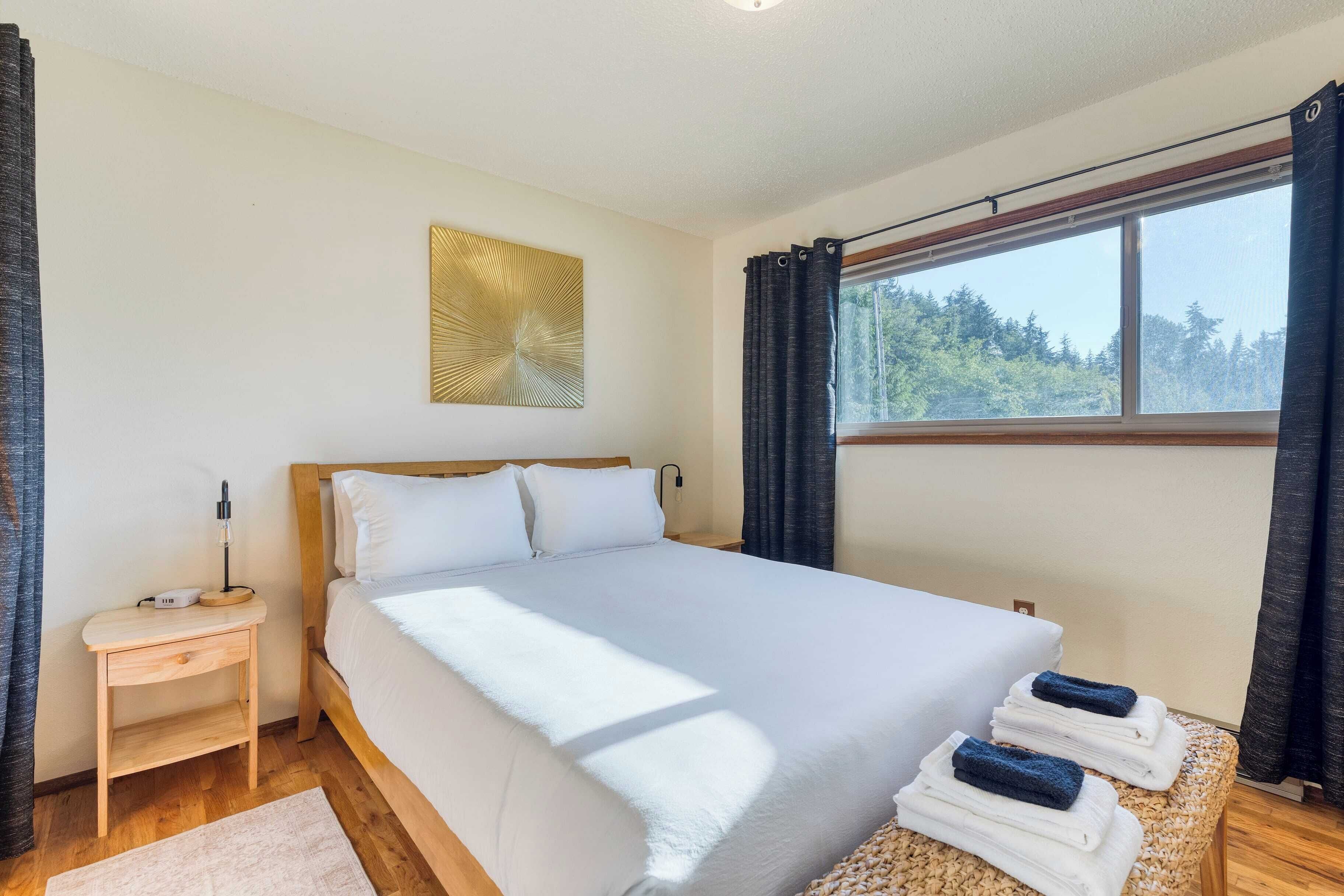 Una cama bien hecha con ropa blanca, una mesita de noche con lámpara y una ventana con cortinas.