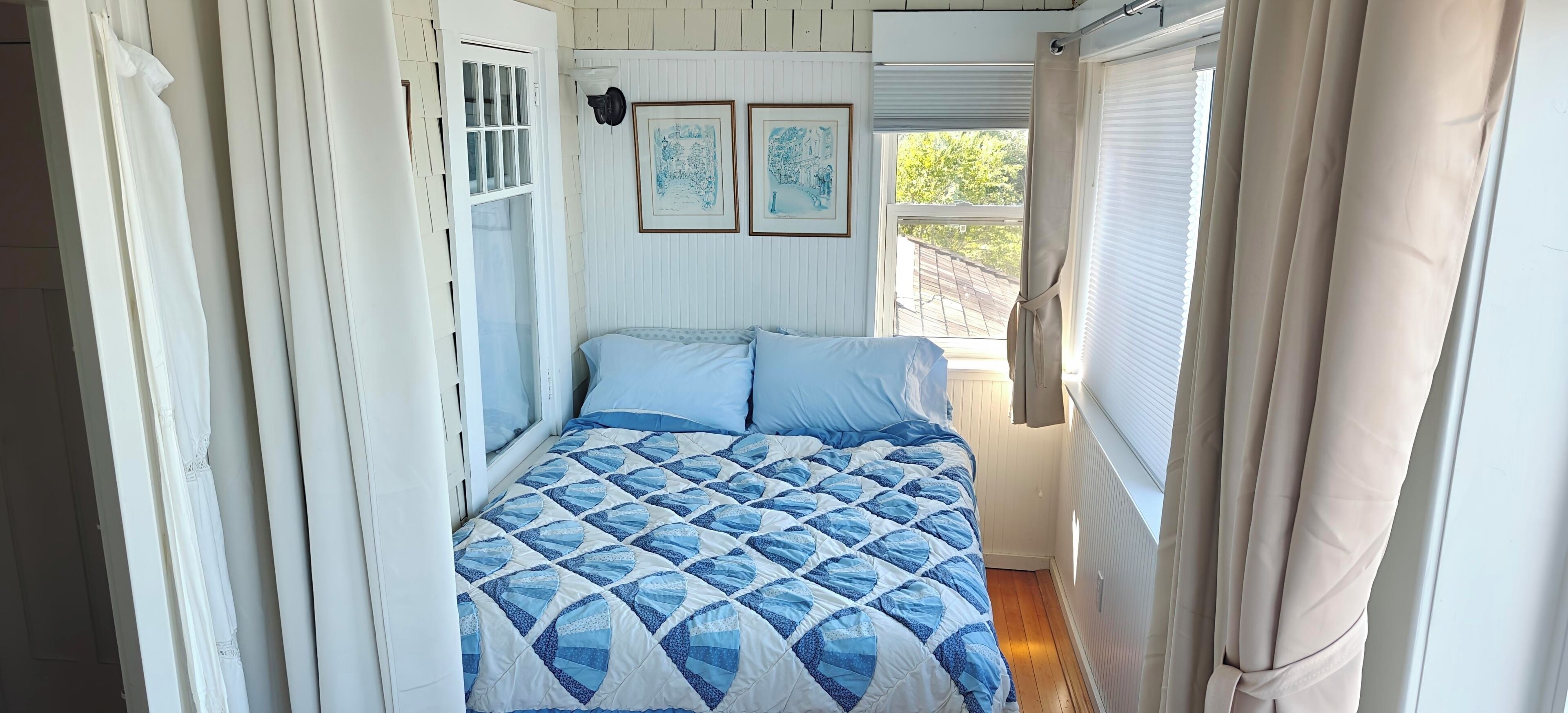 Una habitación pequeña con una cama, una ventana con cortinas y dos cuadros en la pared.