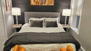 A bed with a grey headboard, two bedside lamps, a cityscape painting above, and a wooden floor.