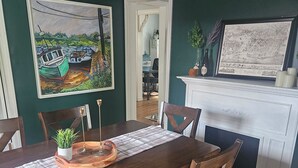 A dining area with a wooden table, chairs, a placemat, and a colorful rug. There is a painting on the wall and a white fireplace with a map above it.